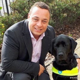 Daniel, who is wearing a suit, is crouching next to a guide dog. They are outside.